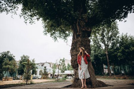 Cute blonde girl in a dress and a red jacket, with a hat on her head, stands near a big tree and looks asideの写真素材
