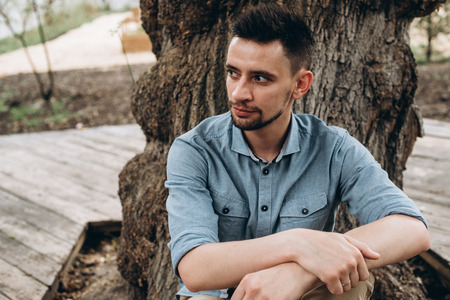 Portrait of a handsome young guy in a blue shirt, who sits near a tree and looks thoughtfully asideの写真素材