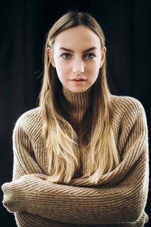 Portrait of a beautiful blue-eyed blonde with long hair in a beige pullover on a black backgroundの写真素材