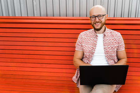 Smiling caucasian confident bearded man with eyeglasses dressed in a stylish wear sitting on a bench outdoors, works remotely using laptop whileの写真素材