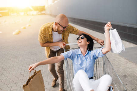 Shopping time. Happy and stylish guy and girl in a sunglasses spend time at the shopping. The smiling girl sits in a cart with paper bag and the guy is push the cart and smiling. Happy time togetherの写真素材