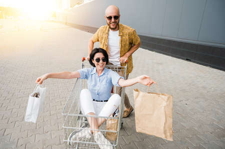 Leisure at the shopping. Shopping together. Happy stylish couple have fun during shopping. The guy push the cart and the girl sits in this cart with paper bags and smilingの写真素材