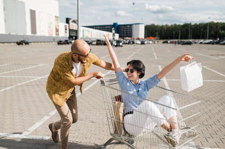 Shopping time. Happy and stylish guy and girl in a sunglasses spend time at the shopping. The smiling girl sits in a cart with paper bag and the guy is push the cart and smiling. Happy time togetherの写真素材