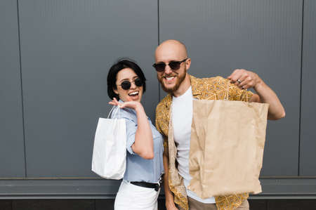 Fashionable guy and girl in sunglasses stand with paper bags after shopping, look at the camera and smile. They are happy to have a good shoppingの写真素材