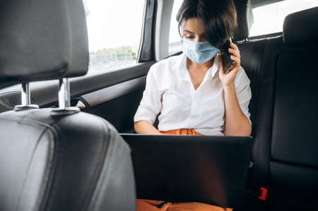 Busy stylish girl sitting in the car in the back seat wearing a medical mask and talks on the phone and working online using a laptopの写真素材