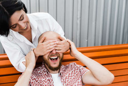 Caucasian couple in love having fun together. The girl stands behind the guy and closes his eyes with her hands, they happy and laughing togetherの写真素材