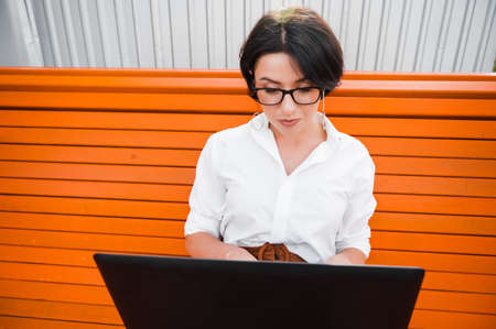 Young attractive business woman wearing eyeglasses dressed in stylish formal clothes works remotely while sitting on a bench outdoors, using a laptopの写真素材