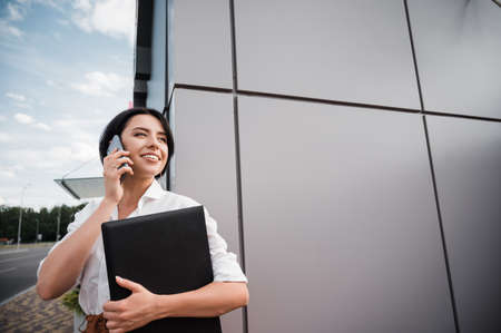 Smiling caucasian business woman dressed in stylish formal wear stands outdoors holding a laptop and...
