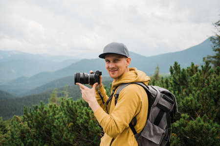 Young male traveler photographer. Attractive man photographer in hiking wear stands in the mountains holding a camera in his hands, smiles and looks at the camera against the backdrop of a mountainsの写真素材