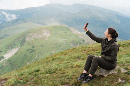 Caucasian happy young woman climbing the top of the mountain and taking a selfie on the phone while sitting on the stone against the background of an amazing mountain landscapeの写真素材