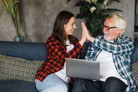 Young adult daughter and gray-haired father are sitting on the couch at home. The girl teaches her...