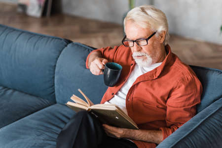 Gray-haired elderly man in eyeglasses sitting at home on the sofa reading a book and holding a cup...
