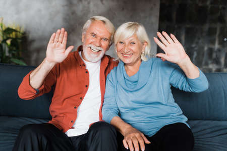 Happy smiling caucasian senior couple sitting at home looking at camera and waving hands during...