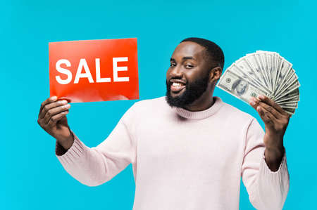 Handsome African American man holds a red "sale" sign and money in his hand standing on a blue background looks at the camera and smilingの写真素材