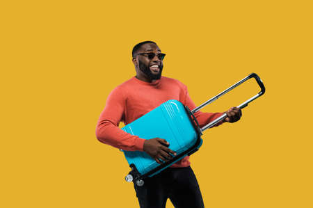 Cheerful African American guy in a sunglasses and in casual wear holds a blue suitcase in his hands standing on isolated orange background. In anticipation of travelの写真素材