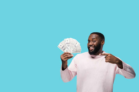 Winning a lottery or time of salary. Cheerful african american man holds bunch of money in his hand enjoying success, standing on isolated blue background and points finger on the moneyの写真素材
