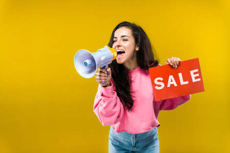 Pretty caucasian young woman emotionally shouts into a megaphone and holds a red "sale" sign in his hand standing on isolated yellow backgroundの写真素材