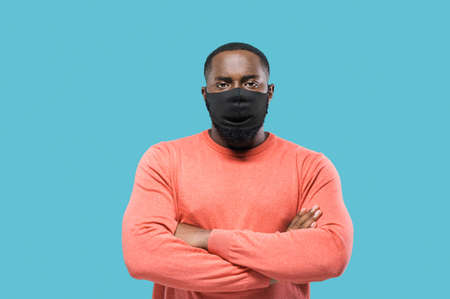 Close-up portrait of african american man in protective black mask because of covid-19 infection or another flu standing against isolated blue background with hands crossedの写真素材