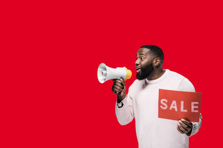 Attractive African American man speaks into a megaphone and holds a red "sale" sign in his hand stand on a red backgroundの写真素材
