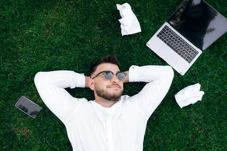 Handsome guy on the grass. Young attractive guy in a white shirt and sunglasses resting from work while lying on the lawn. Nearby lies a laptop, phone and crumpled work papersの写真素材