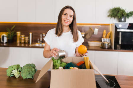 Home food delivery. Young smiling woman is sorting out a box with various vegetables, fruits and other products on the kitchen table which the courier delivered to her homeの写真素材