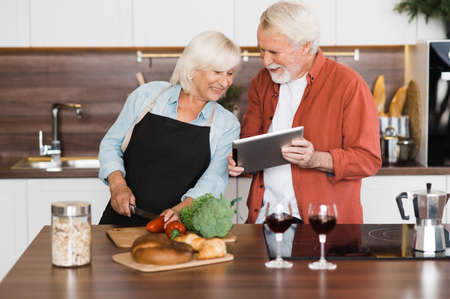 Happy mature couple in the kitchen, wife cooking lunch and her husband shows her something on the...