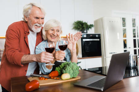 Cheerful elderly gray-haired spouses stand with a glass of wine in the kitchen, communicate and waving hands by video call with family or friends using a laptop in the kitchen at homeの写真素材
