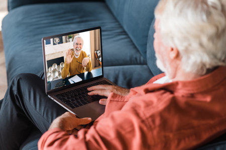 Video call communication. A mature gray-haired man communicates by a video call with his friend, brother or colleague while sitting at home on the couch using laptopの写真素材