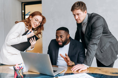 Happy multiracial business people in office using a laptop, look at the screen. Successful coworkers reading good news, get agreement for a project.の写真素材