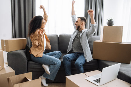 Dreams come true. Overjoyed married multiracial couple buy new apartment, feeling euphoric. Happy caucasian man and african american woman rejoicing, sit on the couch around cardboard boxes, excitedの写真素材