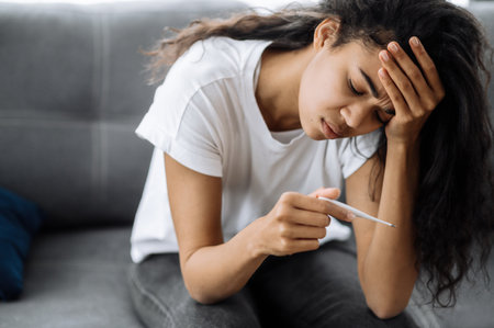 Unhappy sick african american girl sit on couch feel unhealthy, she looks at the thermometer in frustration, ill girl with high fever, she has a headache and needs treatment, flu, quarantineの写真素材