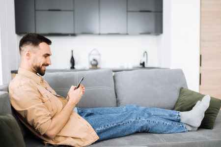 Happy caucasian man sits at the couch, using smartphone. Handsome guy got a dream job or a good news or message. Attractive stylish guy chatting online with friends or familyの写真素材