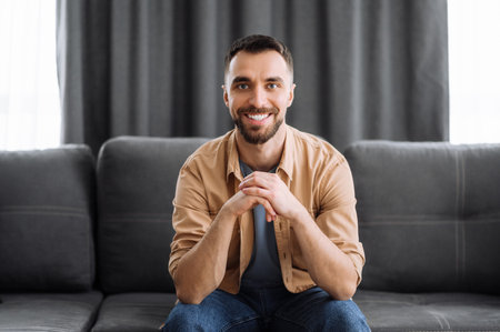 Portrait of an attractive caucasian guy dressed in stylish casual wear. Confident man looks directly at camera with a friendly smile while sitting at the couch in a living roomの写真素材