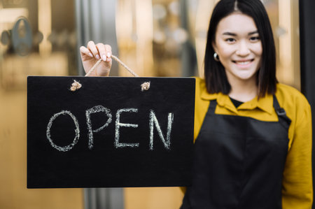 We are open. Defocused satisfied young woman, waitress or owner of a coffee shop or restaurant wearing a black apron stands at the entrance with a signboard OPEN and smiling. Small business conceptの写真素材