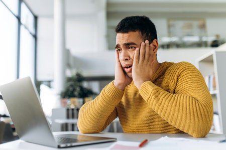 Confused hispanic male freelancer, student or coworker surprised looks at laptop while working or...