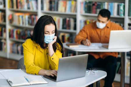 Education during quarantine. Students sit at a distance from each other dressed protective masks on their faces and studying using a laptopの写真素材