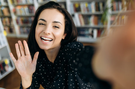 Selfie. Young attractive cheerful caucasian woman takes a selfie on the phone, looks directly at the camera smiling and waving handの写真素材