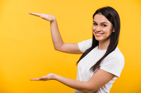 Smiling young caucasian woman in basic white t-shirt showing gesture with hands, standing on isolated orange background and looks at the camera. Copy space for text or advertisingの写真素材