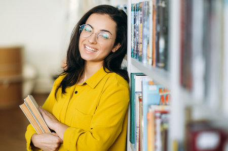 .Portrait of a pretty woman in eyeglasses, standing near bookshelf, looking at the camera. Attractive young female teacher or student hold in arms books, smiling, education concept.の写真素材