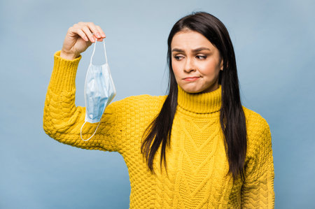 Sad young caucasian woman dressed in yellow sweater hold sterile face mask, standing on isolated blue background .の写真素材