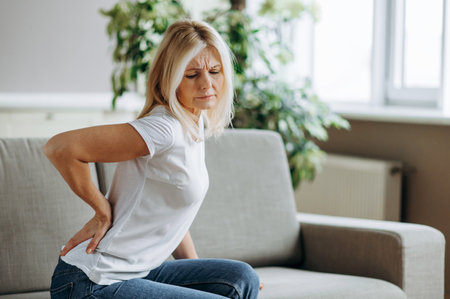 Upset mature woman suffering from backache, unhappy senior blonde sitting on a sofa at living room,...