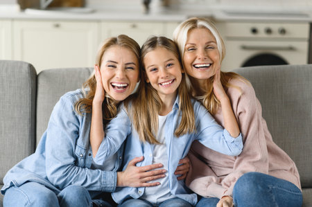 Three female generation. Happy caucasian grandmother, daughter and granddaughter are sitting at home on the couch, hugging, looking at the camera and smiling. Family relationships and valuesの写真素材