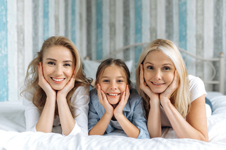 Portrait of three female generation. Happy caucasian grandmother, daughter and granddaughter are lying at home on the couch, looking at the camera and smiling. Family relationships and valuesの写真素材