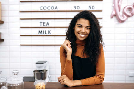 Portrait of a smiling pretty waitress wearing uniform. A young beautiful African American female barista smiles at camera, stands behind the bar counter in coffee shop. Small business ownerの写真素材