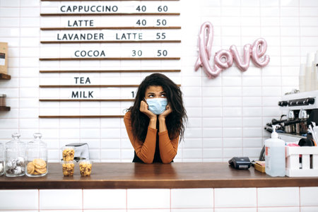 Sad young African American female barista wearing uniform and protective medical mask stands behind the bar counter of coffee shop and waiting for clients. Coffee bar conceptの写真素材
