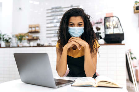 Young African American female small business owner or waitress, wearing medical mask and black apron, studying small business monthly financial statement using laptop, looks at the camera, sitting inの写真素材