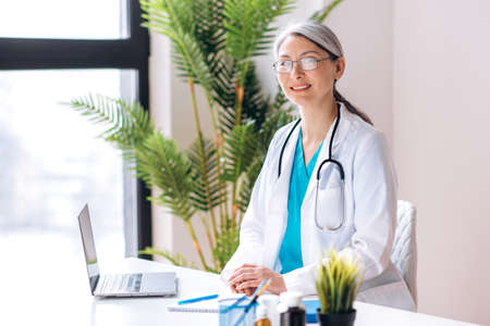 Gray-haired female middle aged general doctor dressed in medical uniform and eyeglasses looks...