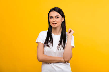Pretty smiling pensive caucasian brunette woman in white basic t-shirt looking pensively to the side biting lip, while standing on isolated orange background, thinking about ideaの写真素材