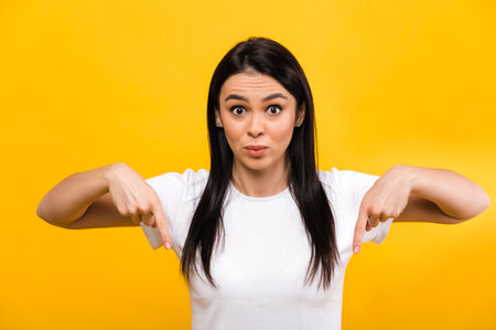 Photo of surprised excited cute caucasian girl pointing fingers down, standing on isolated orange background, with shocked face and looks at the cameraの写真素材