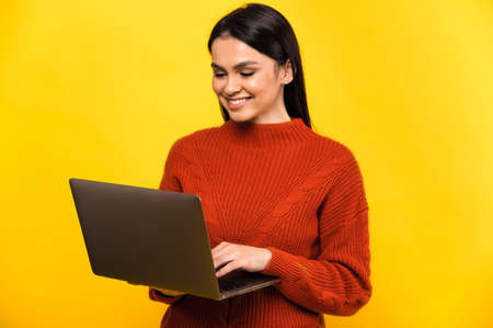 Attractive young caucasian smiling woman in casual sweater, stands on isolated yellow background and working online on her laptop, replies to email, types a messageの写真素材
